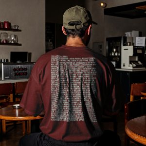 Uomo di spalle seduto in un bar con t-shirt bordeaux e lungo manifesto tipografico in verde acqua stampato sulla schiena