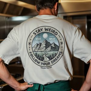 Uomo di spalle con t-shirt chiara e grande grafica tonda “Stay Weird Follow Your Dreams” con montagne, luna e bosco sul retro