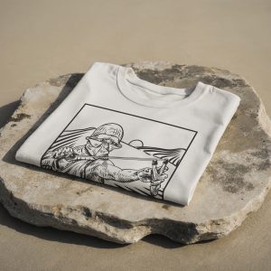 T-shirt color panna piegata con illustrazione in bianco e nero di un ragazzo con casco che tende una fionda, appoggiata su una grande pietra chiara